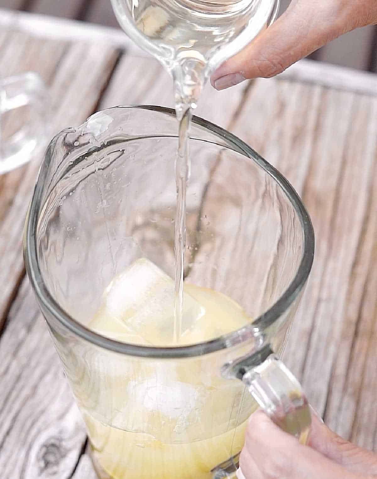 Pouring syrup into a pitcher with lemon juice and ice cubes. Grey wooden surface.