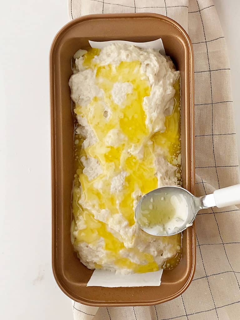 Melted butter being spooned on beer bread batter. Copper loaf pan, beige towel. White surface.