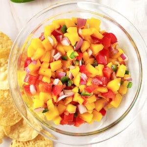 A glass bowl with mango peach salsa. White surface and cloth. Corn chips.