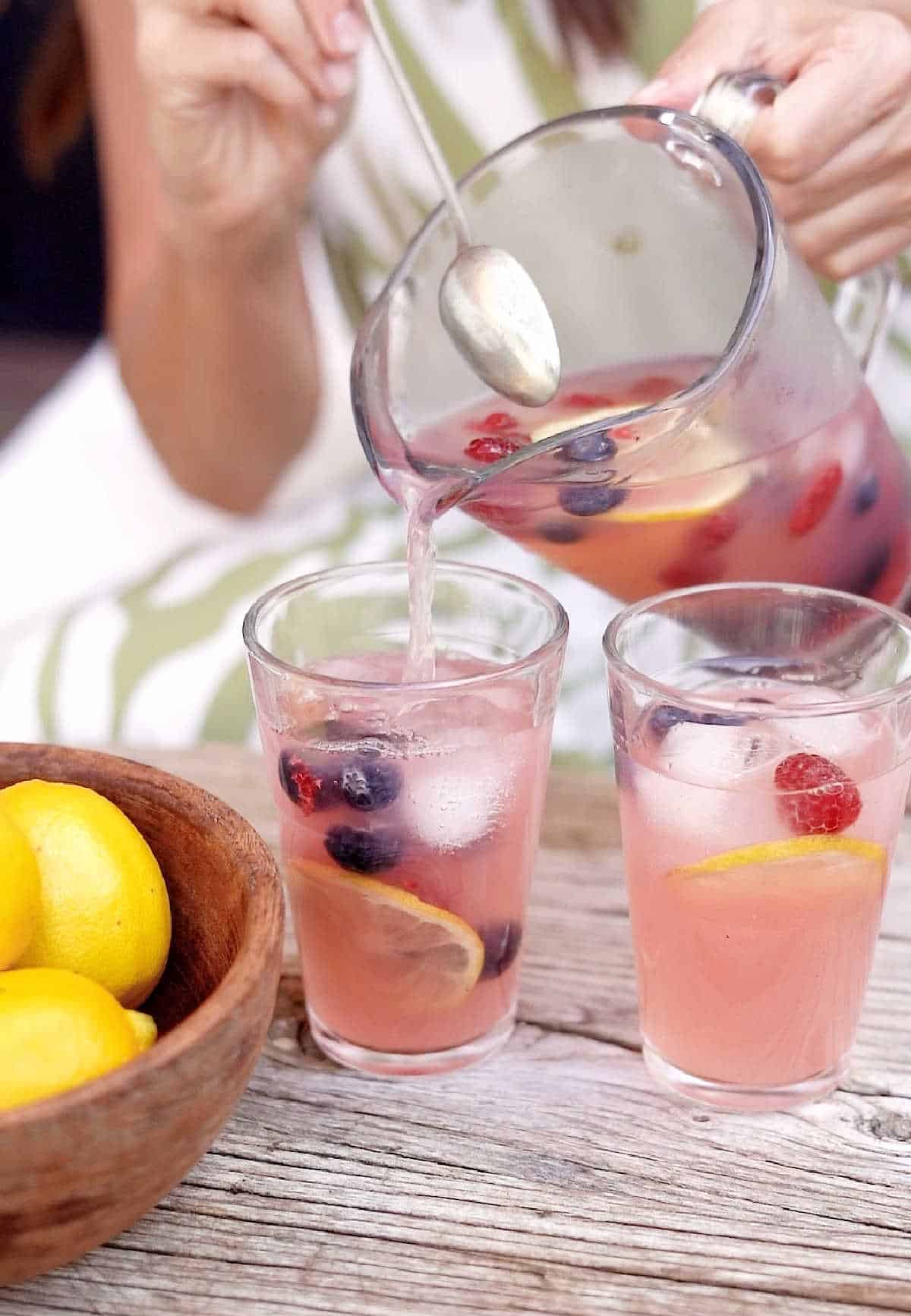 Serving pink lemonade from a pitcher into tall glasses. Wooden table, bowl with lemons. 
