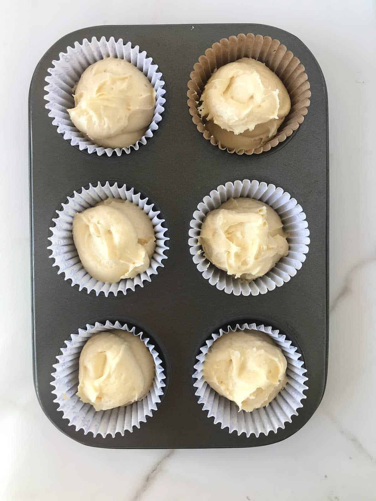 Vanilla cake batter in paper liners in a dark muffin tin. White marbled surface. 