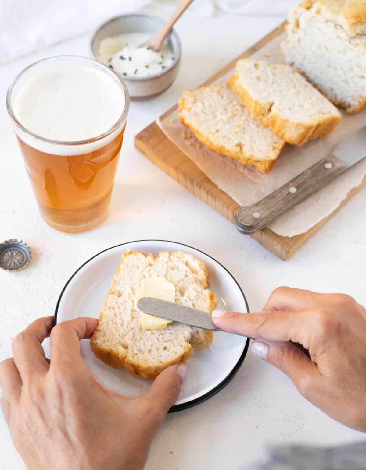 Buttering a slice of beer bread on a white plate. A beer glass, wooden board with loaf of bread, white surface. 