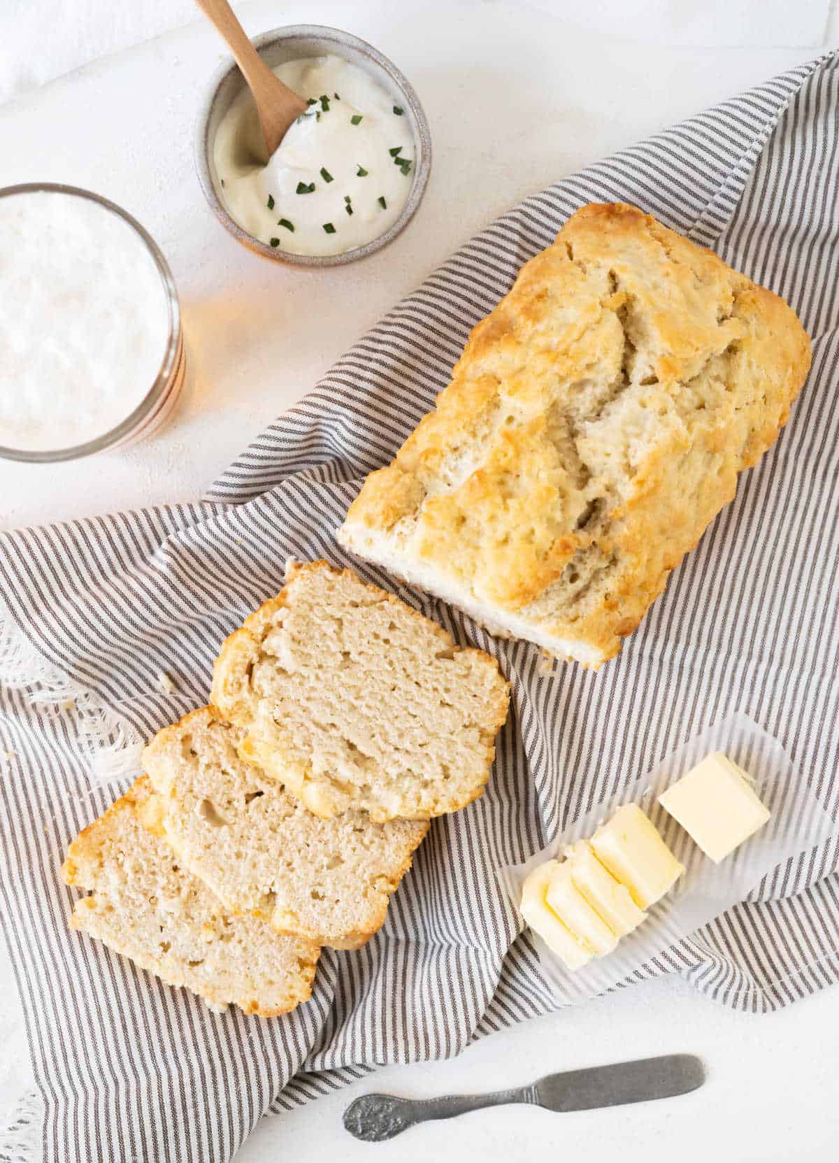Beer bread with slices on a striped towel, butter slices, small bowls. White marble surface.