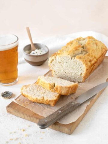 Wooden board with parchment paper on white marble with loaf of beer bread, whole and sliced. A knife, glass of beer and pinkish background.