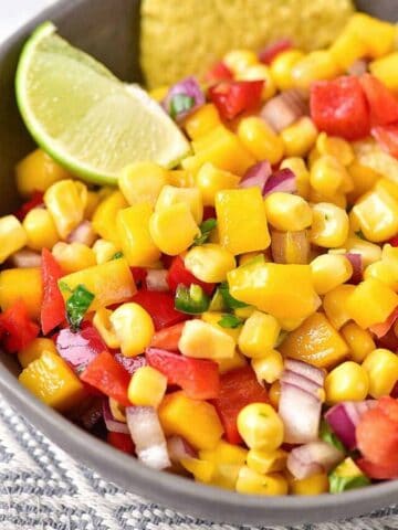 Close up of mango corn salsa in a grey bowl with lime wedge and chips.