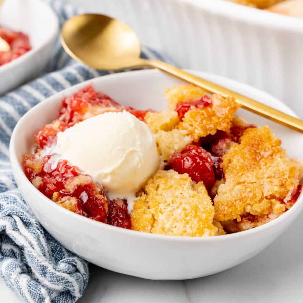 A white bowl with cherry dump cake and scoop of ice cream. Blue striped cloth and gold spoon.