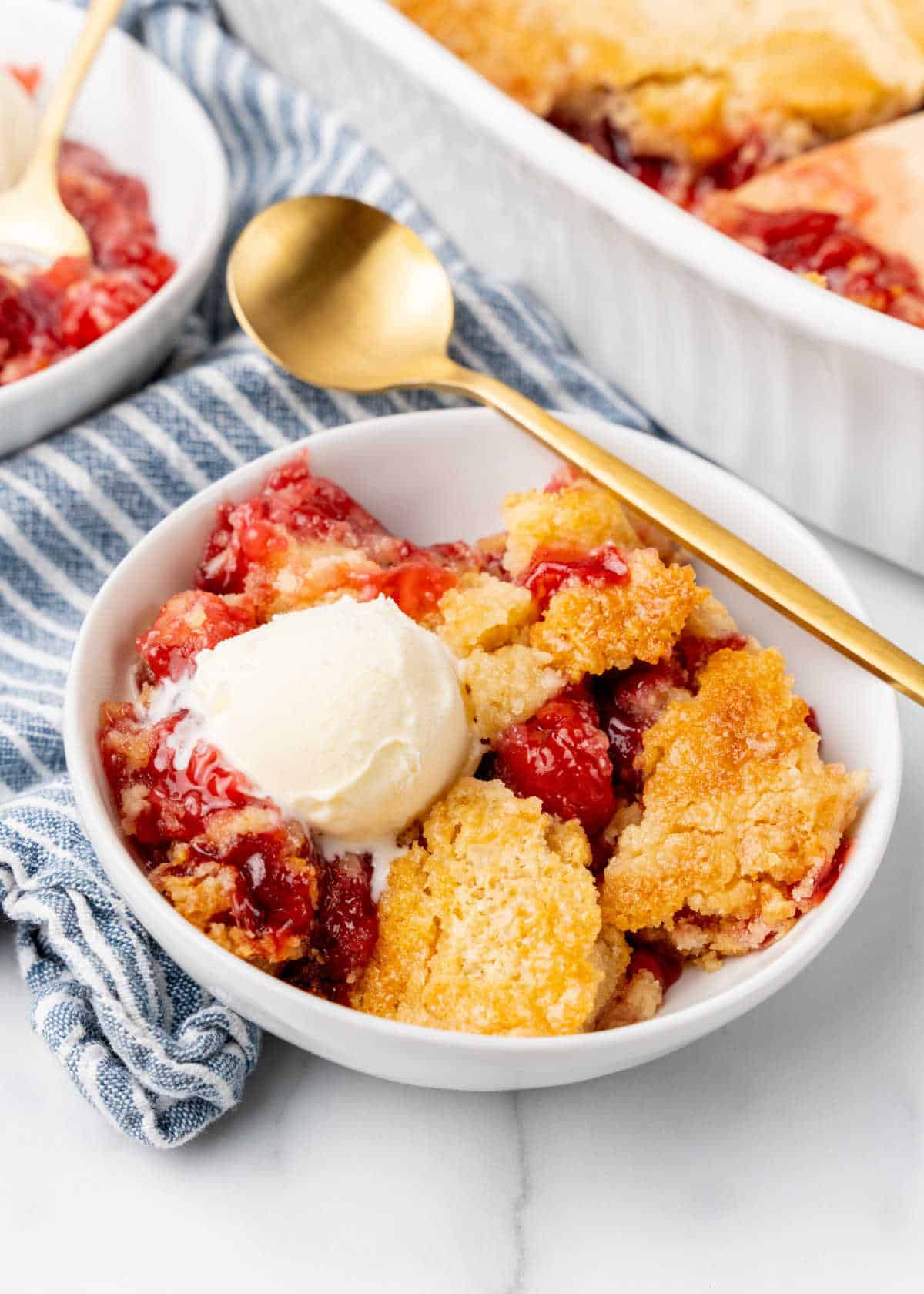 Serving of cherry dump cake with ice cream in a white bowl. A gold spoon, white marble surface, striped blue white cloth.