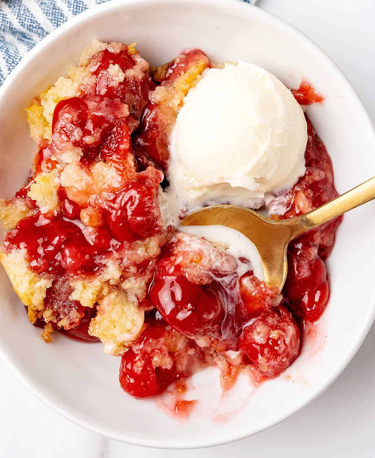 Close up image of cherry dump cake served with ice cream in a white bowl with a gold spoon.