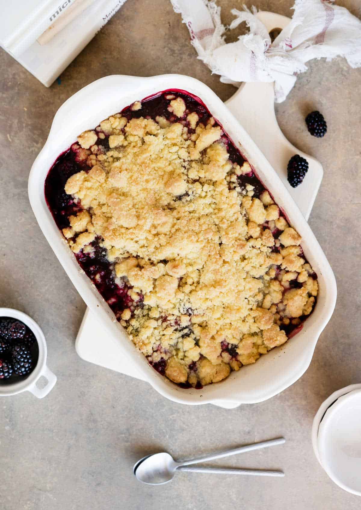 Top view of white rectangular dish with blackberry dump cake on a brownish surface. Bowls, spoons, loose blackberries around.