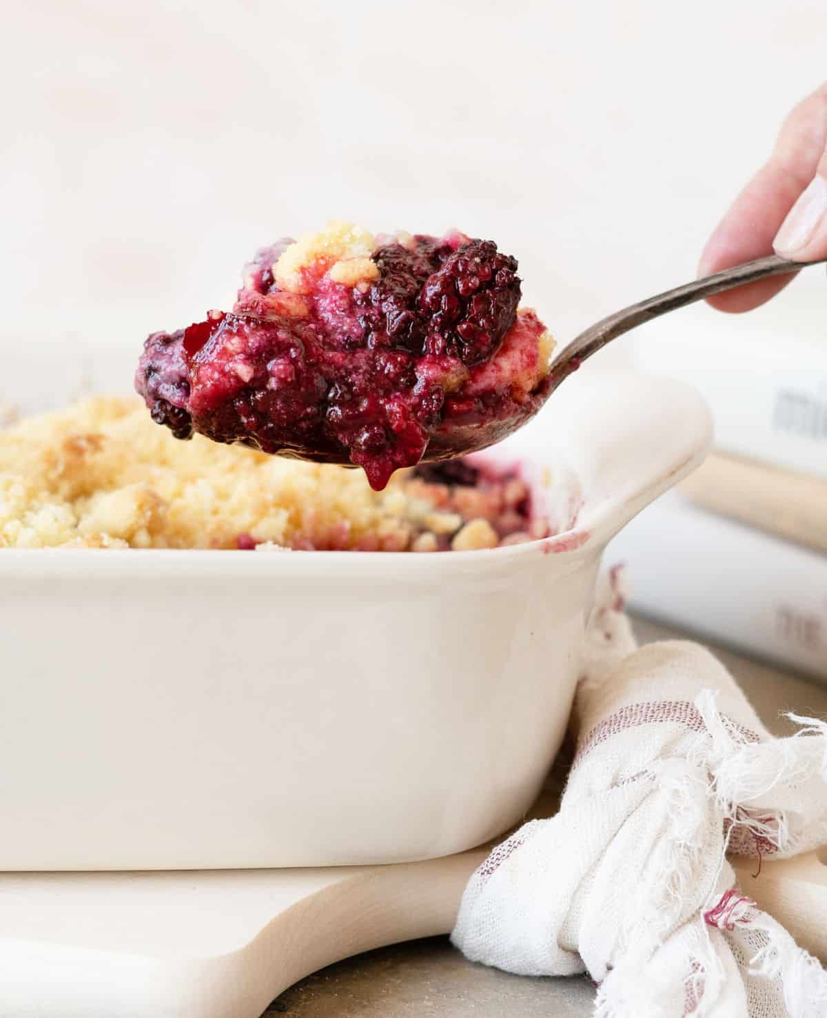 Spoon with blackberry dump cake. White baking dish with kitchen towel on a light peach colored background.