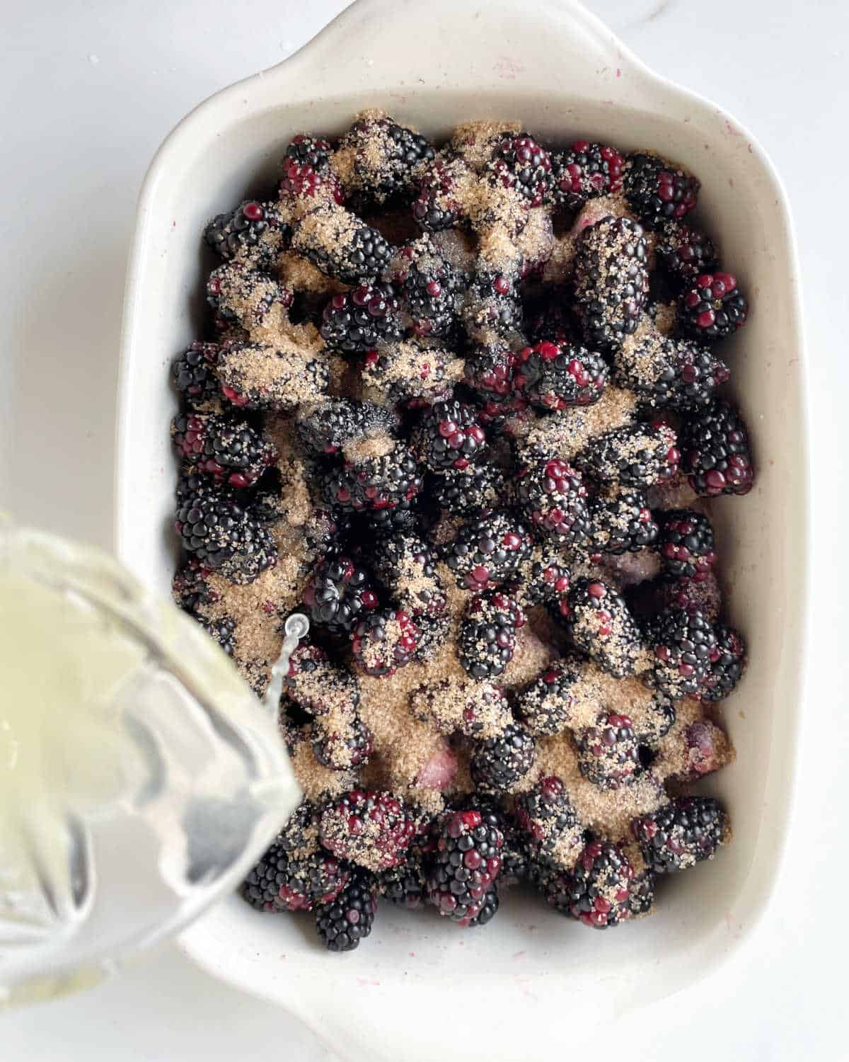 Adding lemon juice to blackberries and brown sugar in a cream colored dish on a white surface.