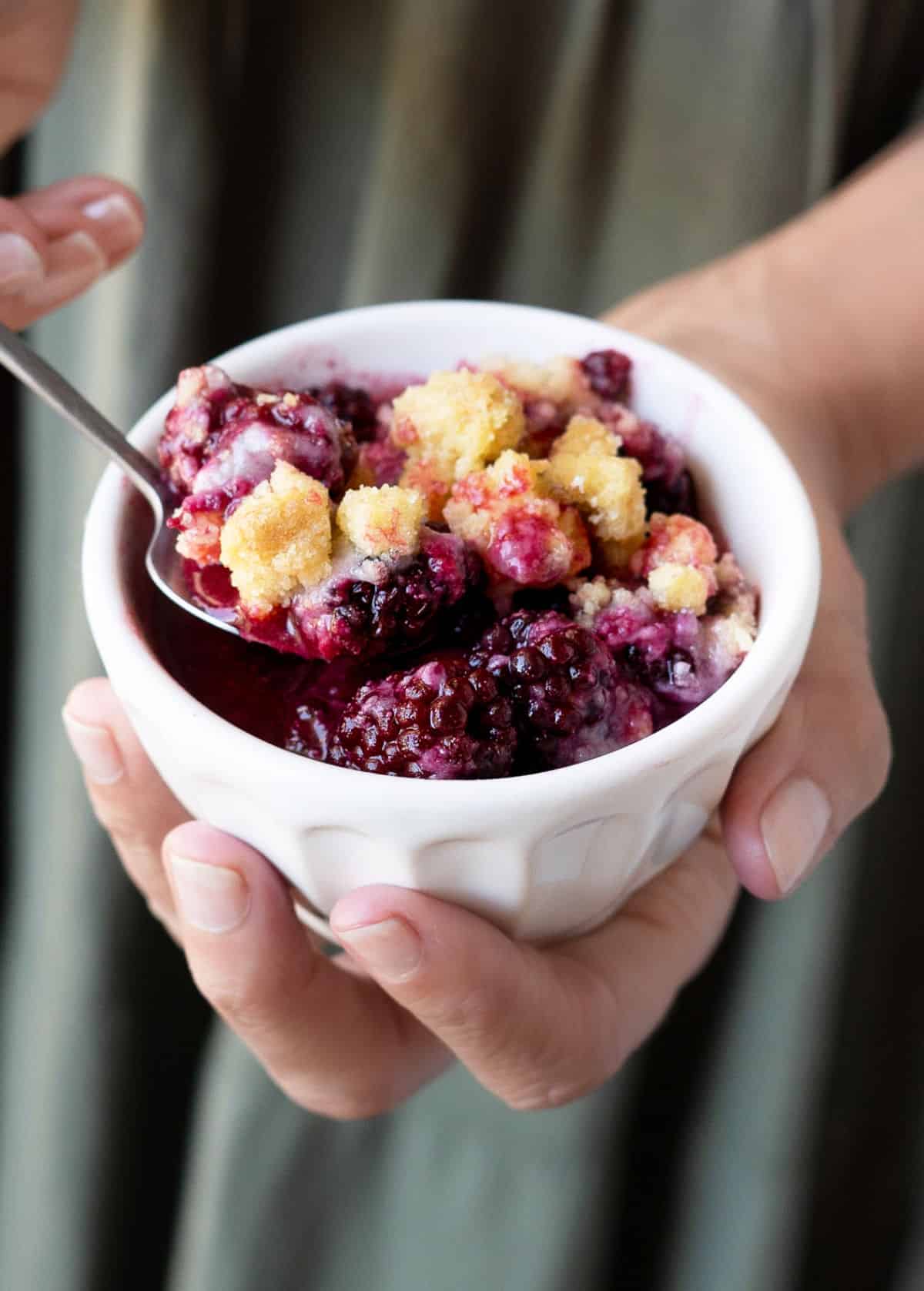 Blackberry dump cake serving in a white bowl with a spoon being held by someone in a green dress. 
