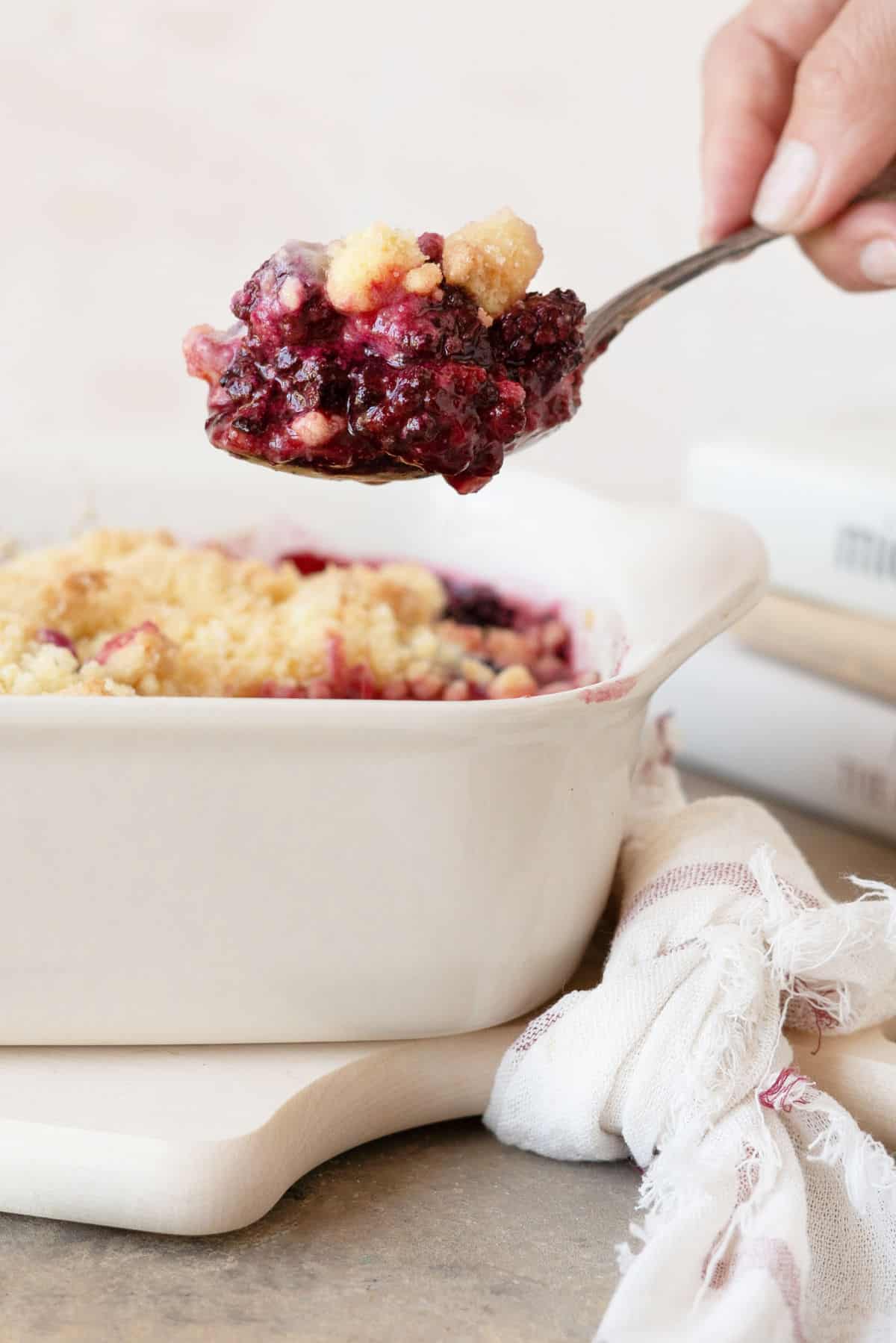 Hand lifting blackberry dump cake with a spoon. White dish on a board, a kitchen towel, peach colored background. 