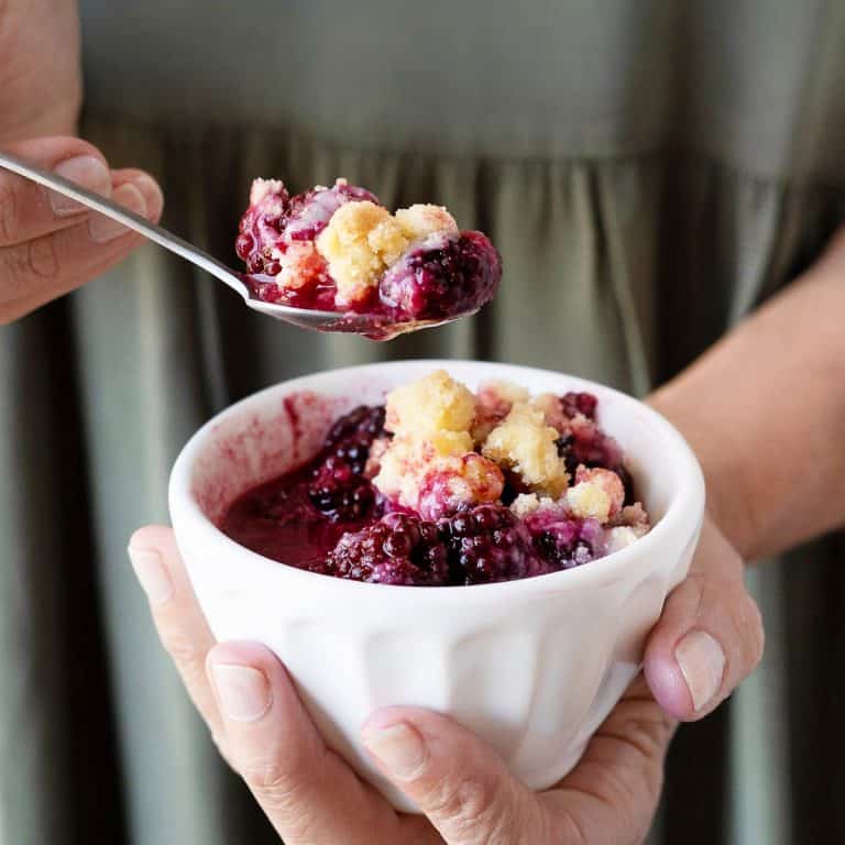 White bowl with berry dump cake being held by person in a green dress lifting a full spoon.
