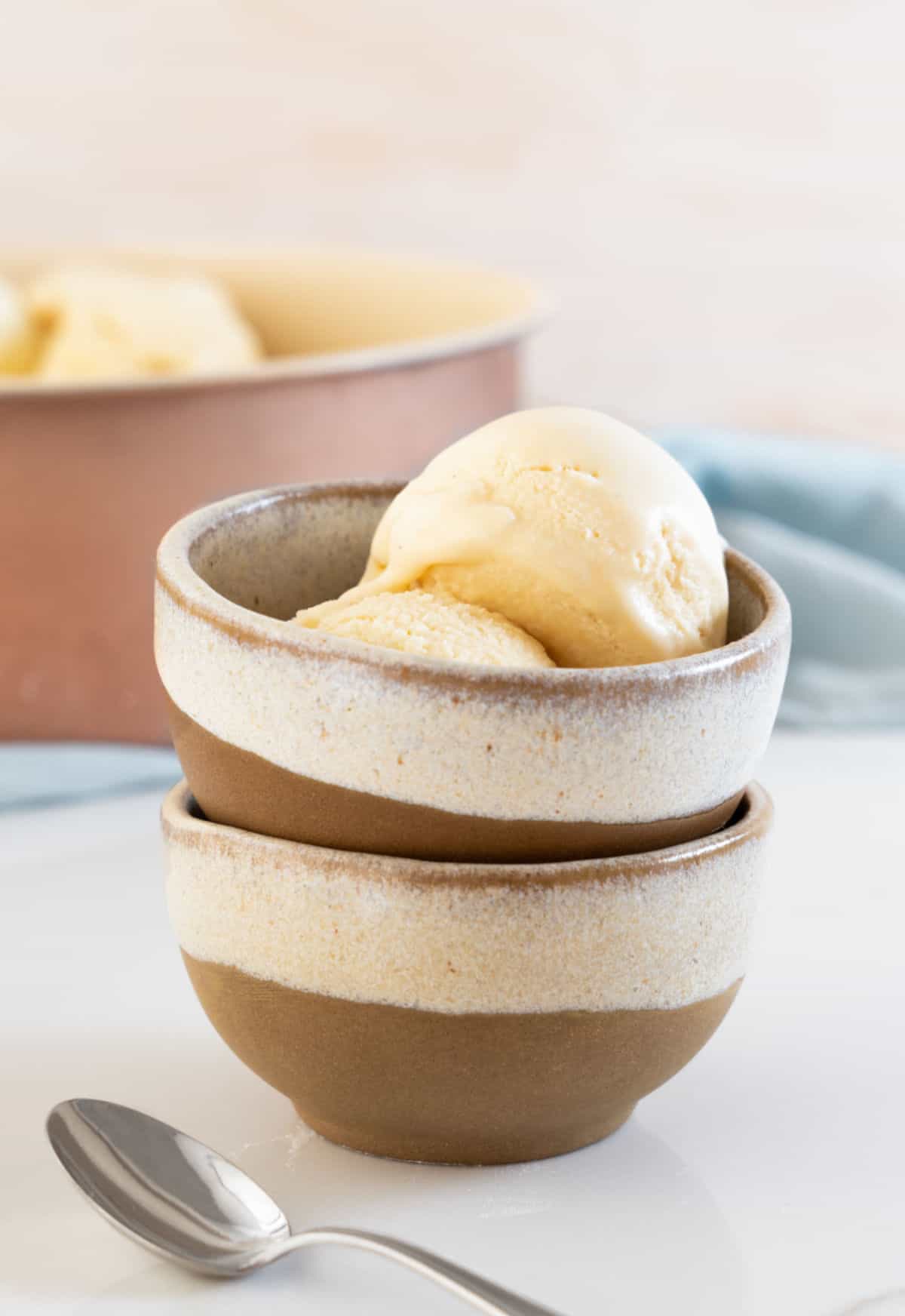 Two stacked brown beige bowls with scoops of vanilla ice cream. White surface, pink background, light blue cloth. 