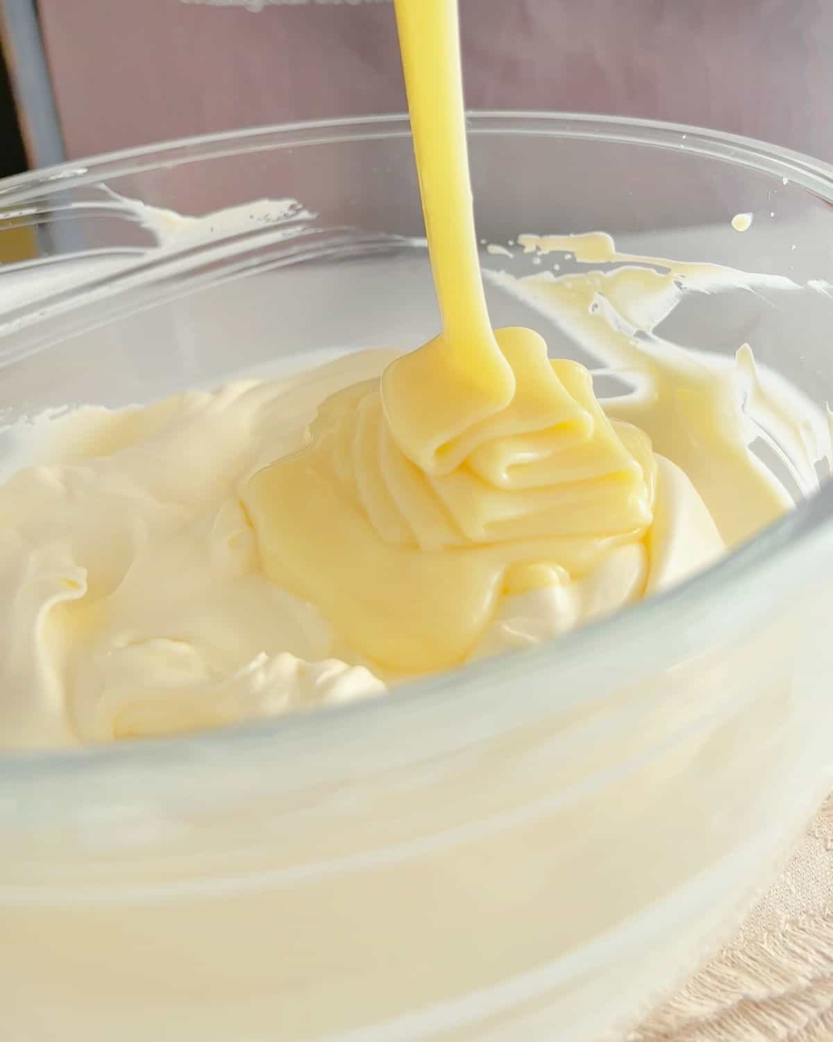 Pouring condensed milk on whipped cream in a glass bowl.