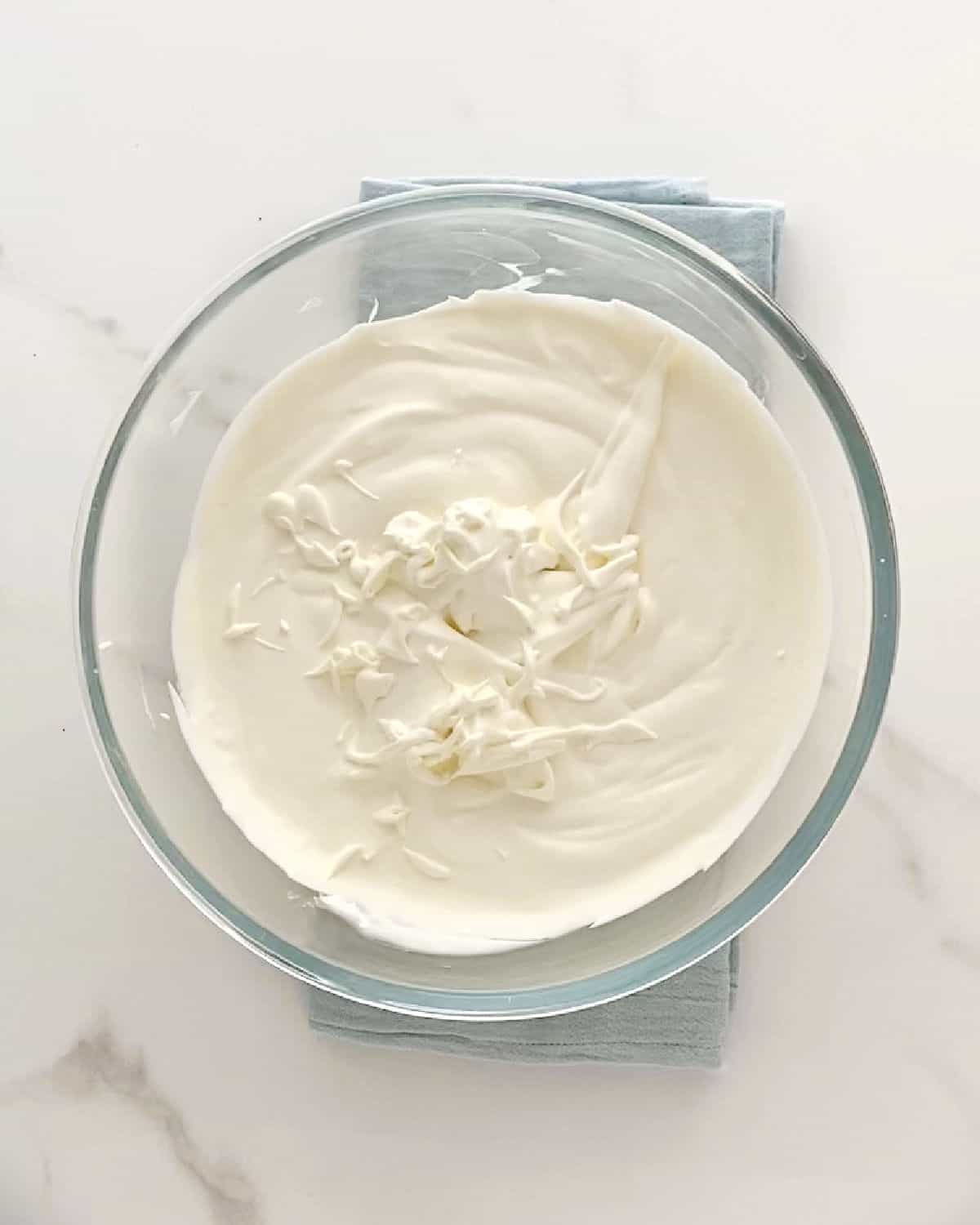 Glass bowl with whipped cream on a light blue cloth on a white marble surface.