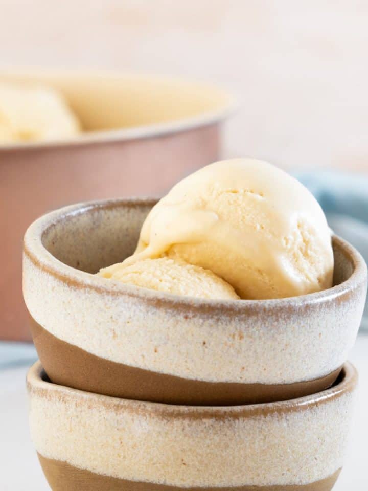 Scoops of vanilla ice cream in a stack of two beige brown bowls. Metal pan and light blue cloth in the pink background.