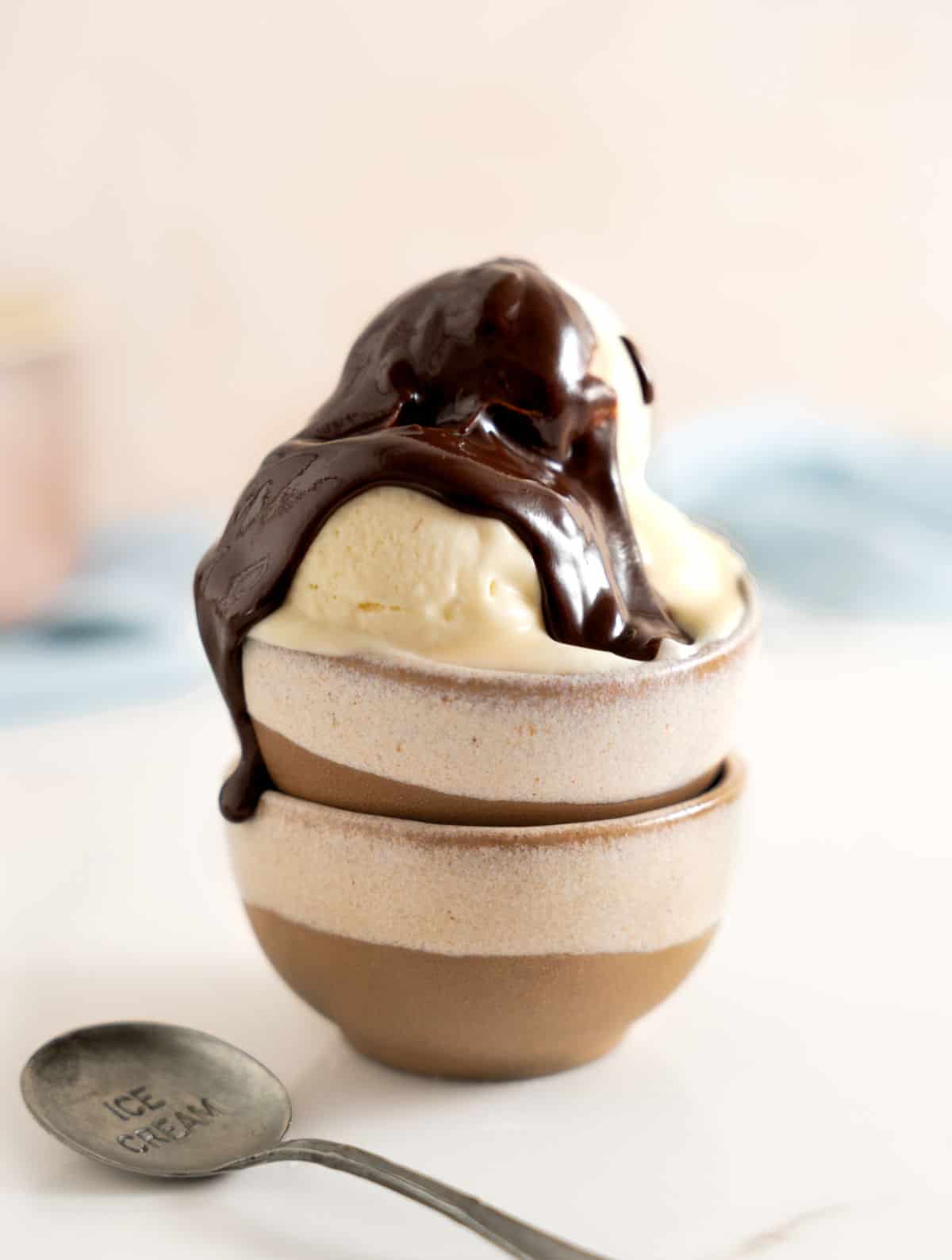 Chocolate sauce on vanilla ice cream on beige brown bowls with a spoon. White surface, pink blue background.