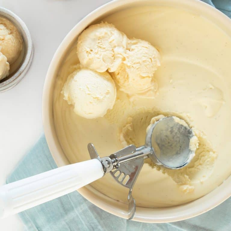 Close up of vanilla ice cream in a metal round pan with a white spoon, several scoops. White and light blue surface.