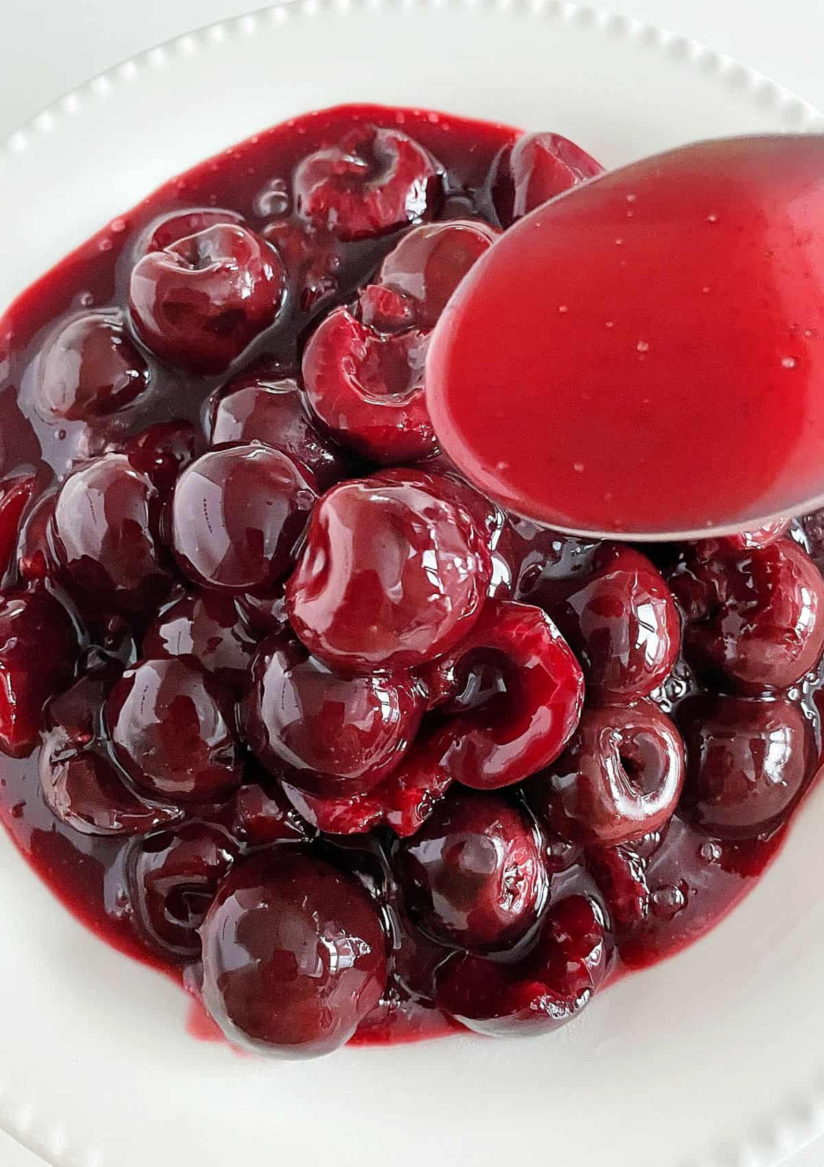 Cherry syrup dripping from spoon over white bowl of cherry sauce.
