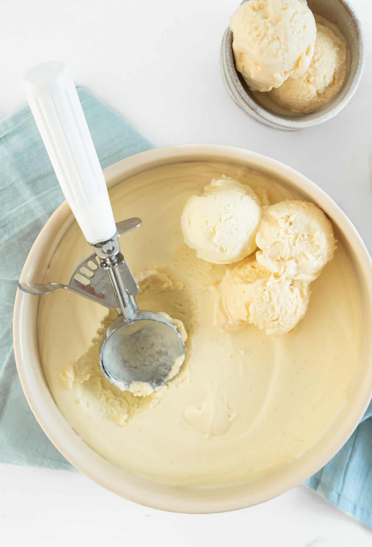 Round metal beige pan with vanilla ice cream. Scoops with white serving spoon. Light blue cloth on a white surface. 