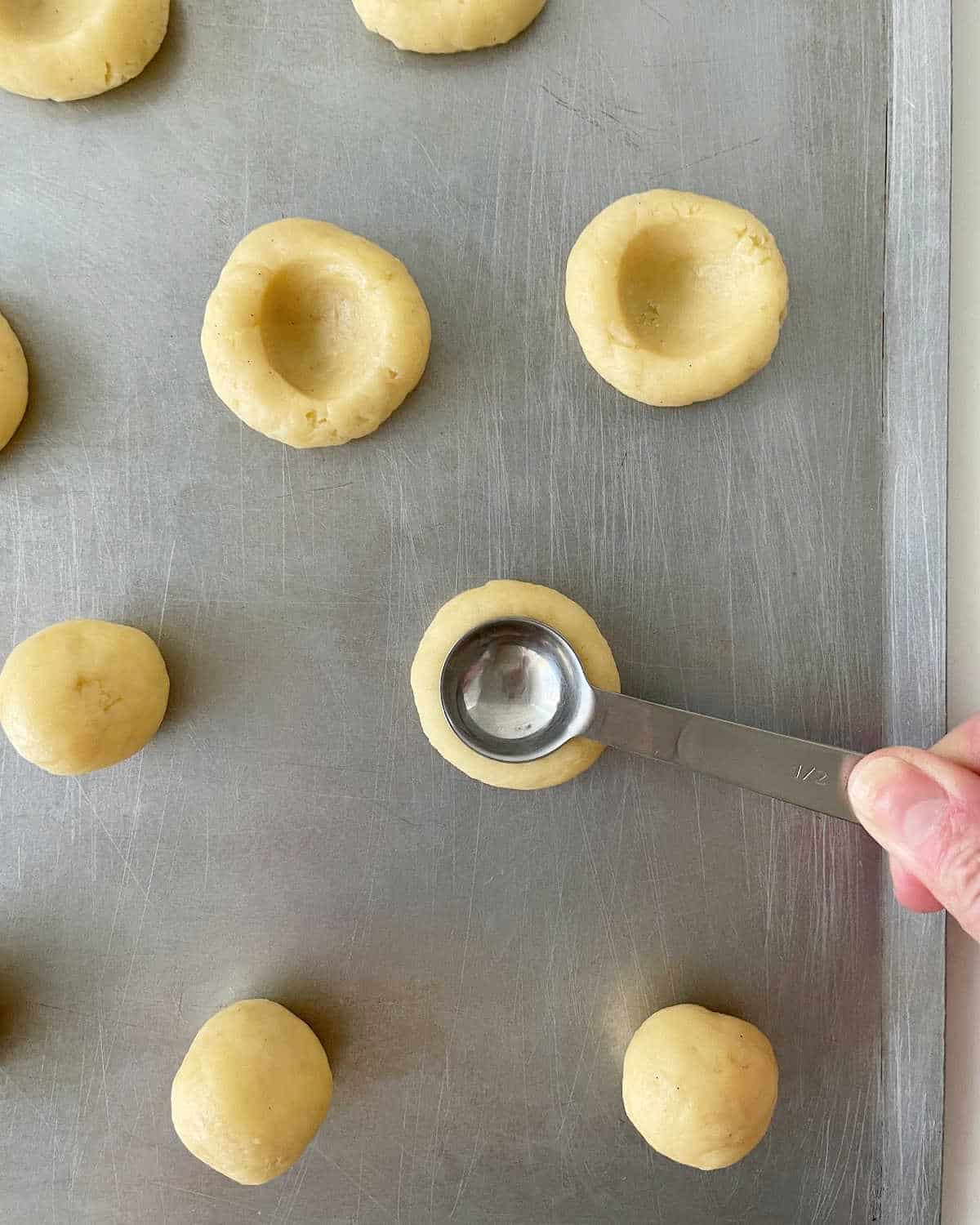 Making thumbprint indentation with a measuring spoon on cookie dough balls on a baking sheet.