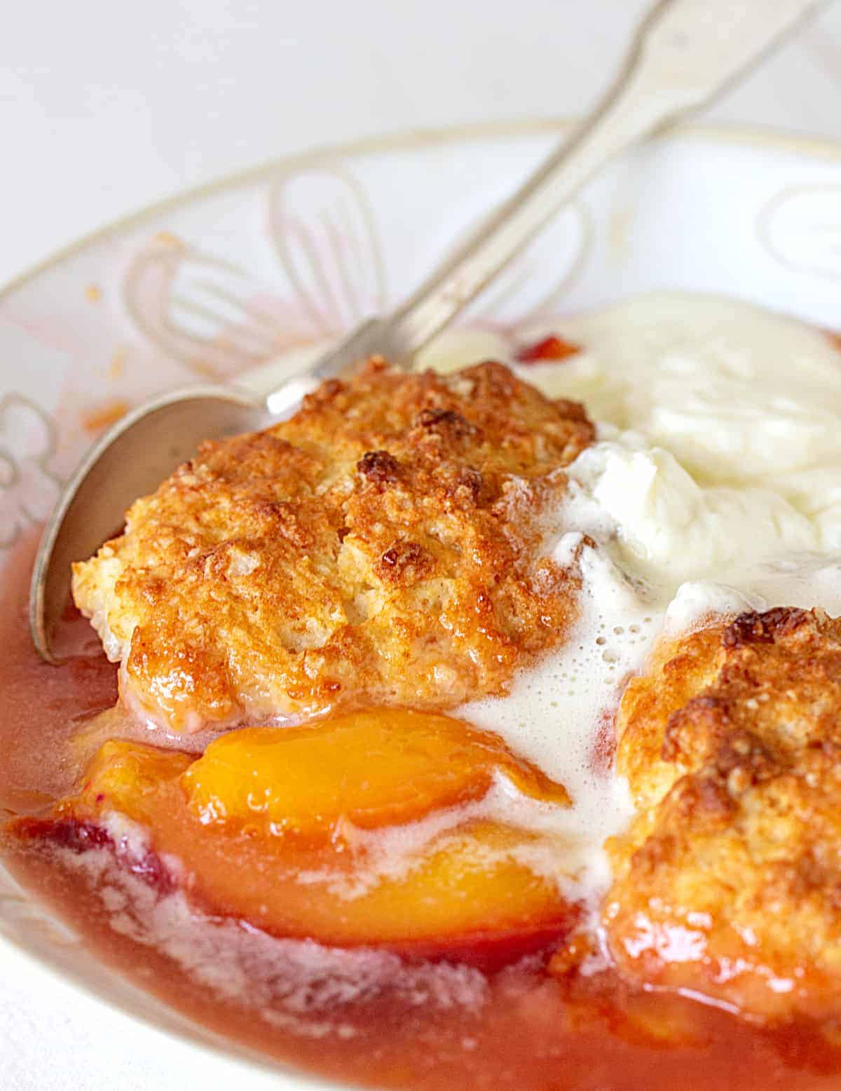 White bowl with juicy peach cobbler, scoop of ice cream, silver spoon.