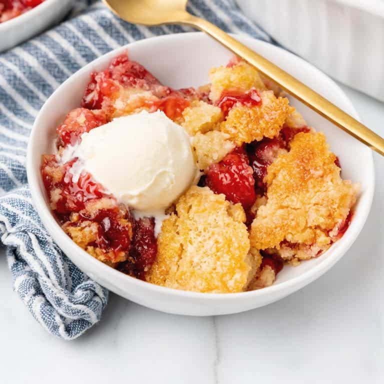 Striped blue white kitchen towel with white bowl containing serving of cherry cobbler with ice cream. A gold spoon, white marble surface.