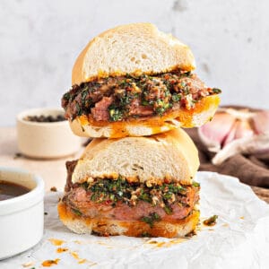 Close up stack of chimichurri steak sandwich on white parchment. Bowls with sauce, light gray background.