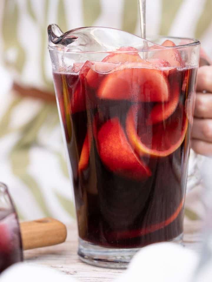 Close up of glass pitcher with red wine sangria stirred by person in a green white dress.