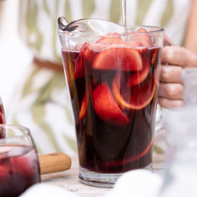 Close up of glass pitcher with red wine sangria stirred by person in a green white dress.