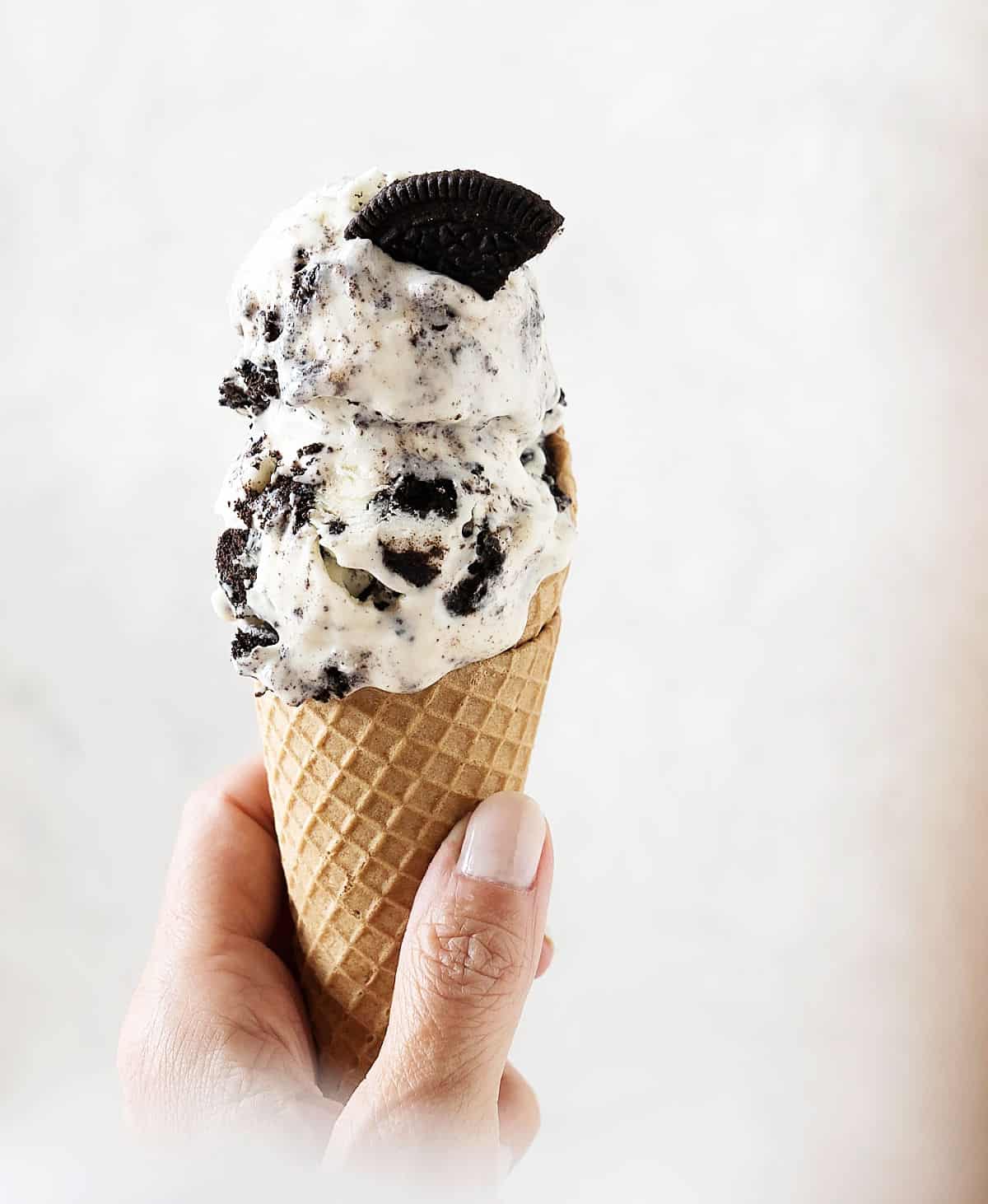 Hand holding a waffle cone with scoops of Oreo ice cream. Light grey background. 
