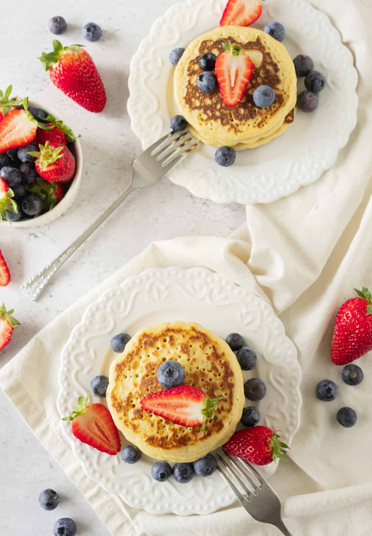 Two white plates with stack of cornmeal pancakes with white berries on a white surface. Top view.