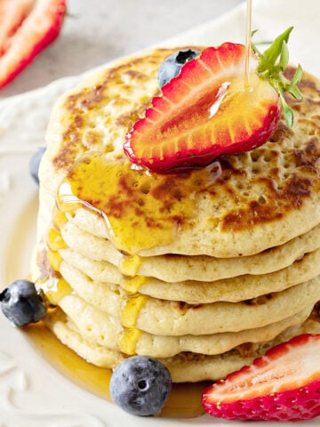 Several cornmeal pancakes stacked on a white plate. Berries and syrup.