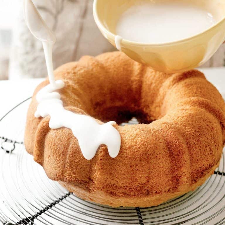 Drizzling a bundt cake on a wire rack with powdered sugar glaze in a yellow bowl.