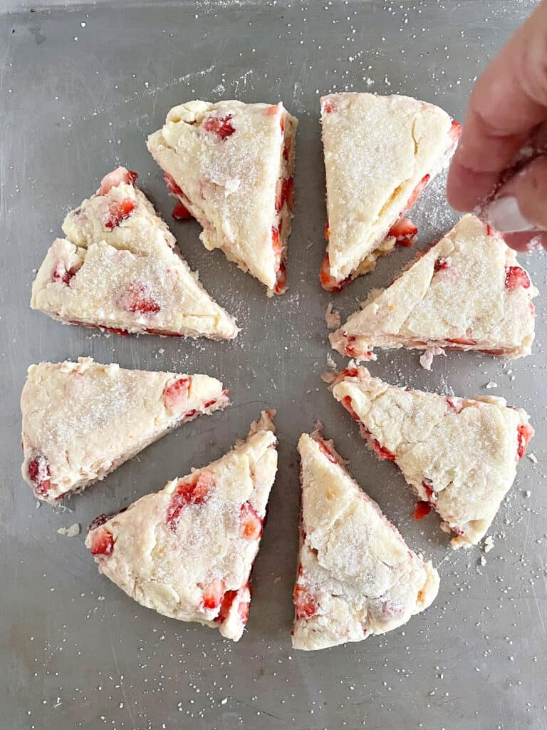 Sprinkling sugar on unbaked strawberry scones triangles on a metal tray.