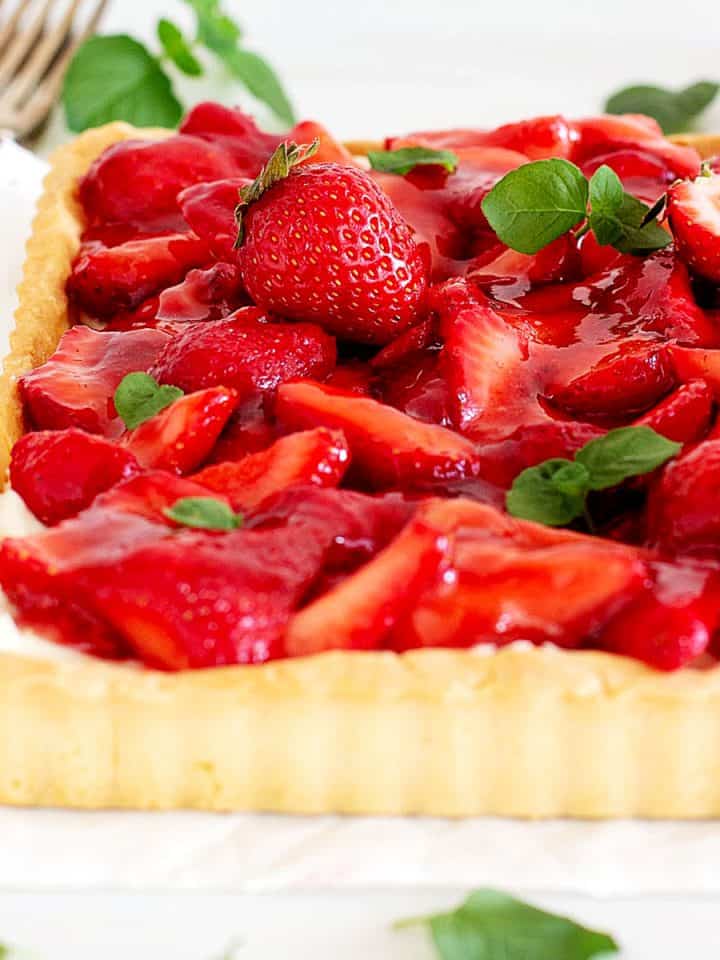 Partial view of square fresh strawberry tart on a white surface with mint leaves.