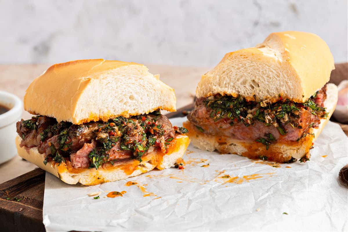 Halved steak chimichurri sandwich on white paper.