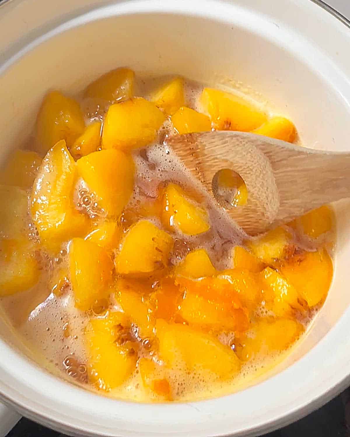Wooden spoon mixing a peach compote simmering in a white saucepan.
