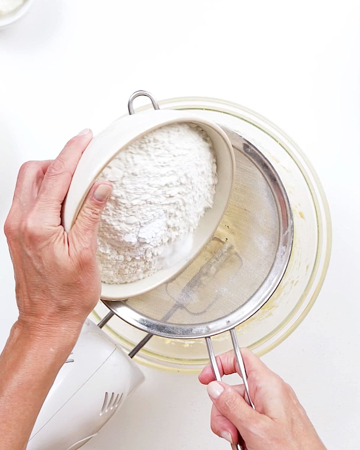 Sifting flour over glass bowl with cake batter. White surface.