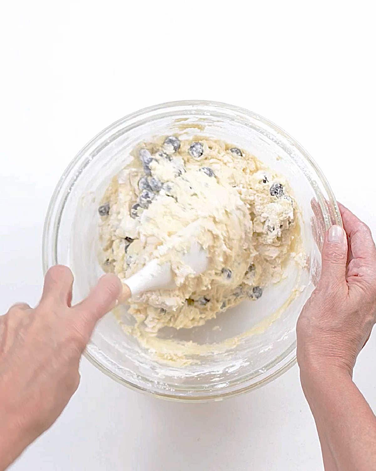 Mixing blueberry cake batter in a glass bowl with a white spatula. White surface.