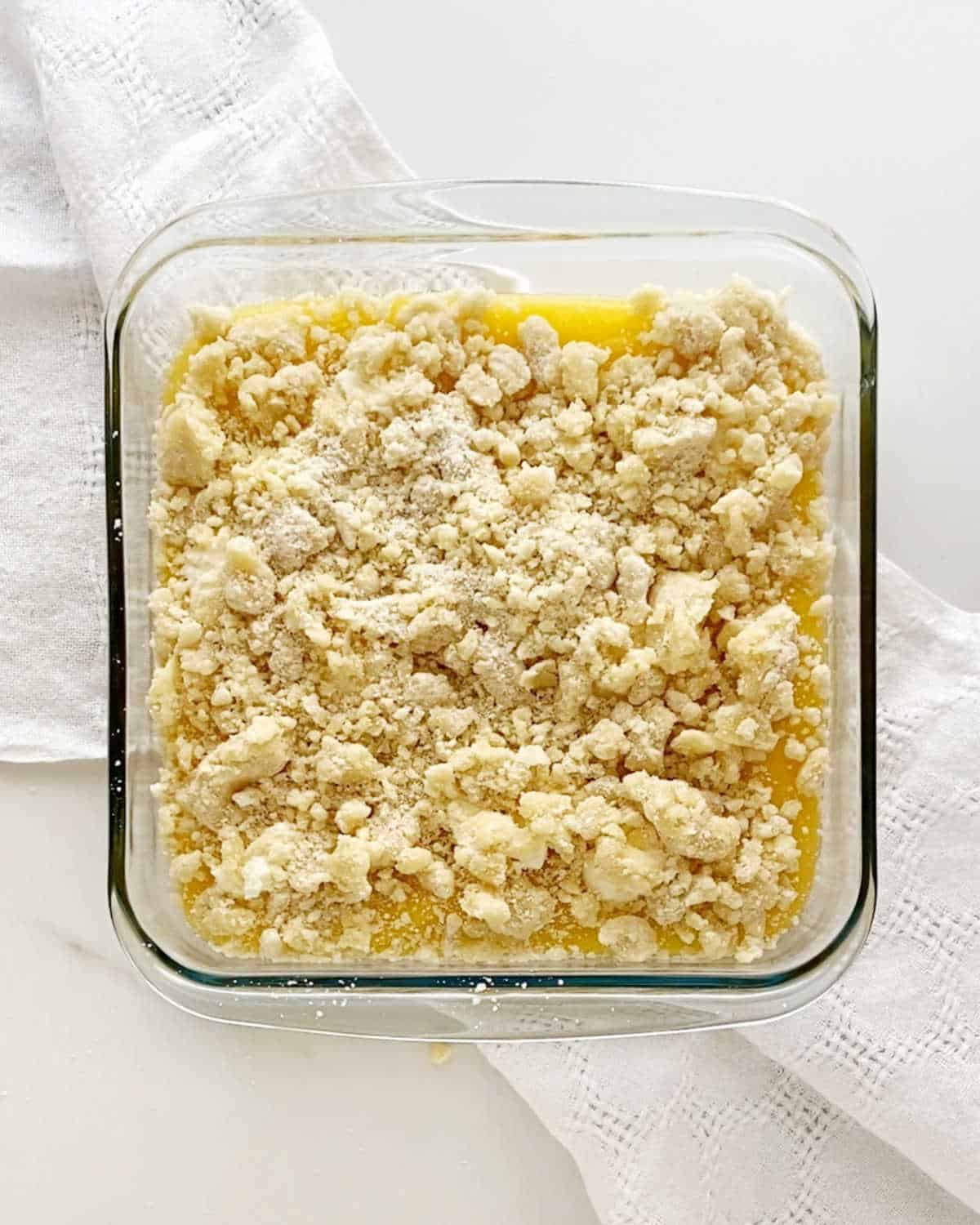 Crumble topped lemon dump cake before baking in a glass square baking dish. White cloth and white marble surface.
