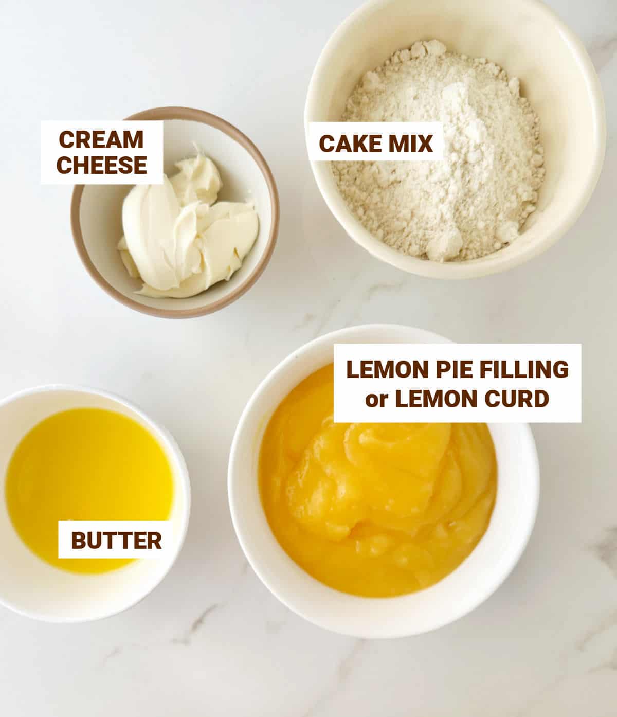 Bowls on a white surface with ingredients for lemon dump cake including cream cheese, lemon curd, butter and cake mix.