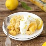 Serving of lemon dump cake with cream on white plate with a silver spoon. Wooden surface with whole lemon in the background.