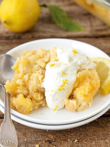 Serving of lemon dump cake with cream on white plate with a silver spoon. Wooden surface with whole lemon in the background.