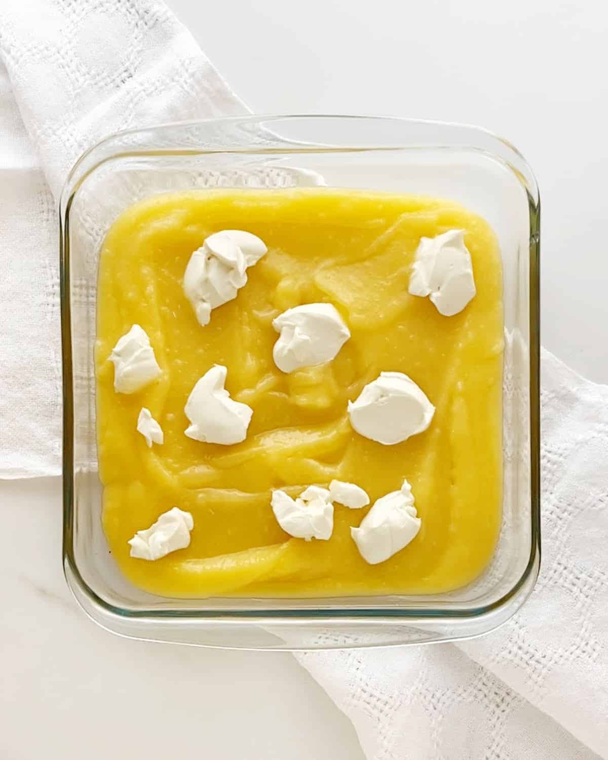 Glass square dish with lemon curd and cream cheese dollops on a white cloth and surface.