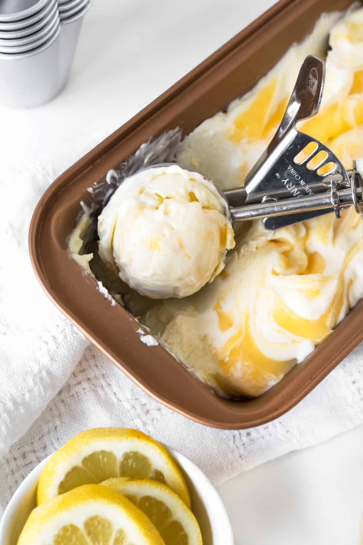 Copper loaf pan with lemon swirl ice cream, a scoop, white surface, lemon slices.