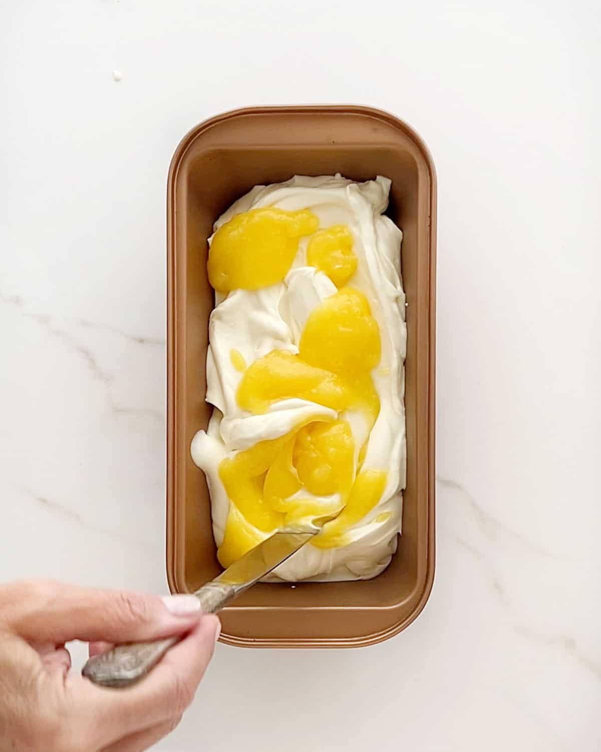 Swirling lemon curd into ice cream in a copper loaf pan. White marble surface.