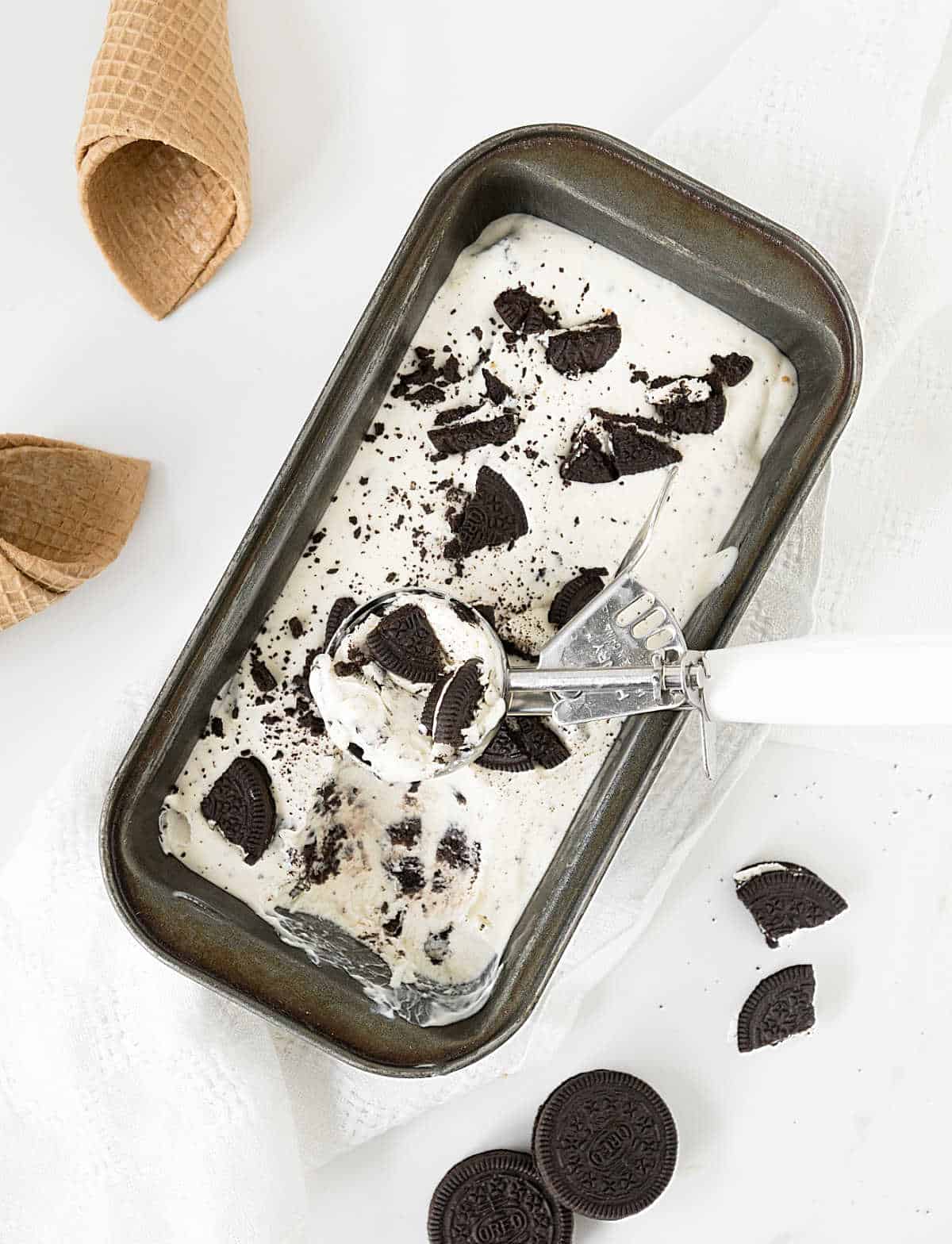 Loaf pan with oreo ice cream, an ice cream scoop, loose cookies, cones. White surface.